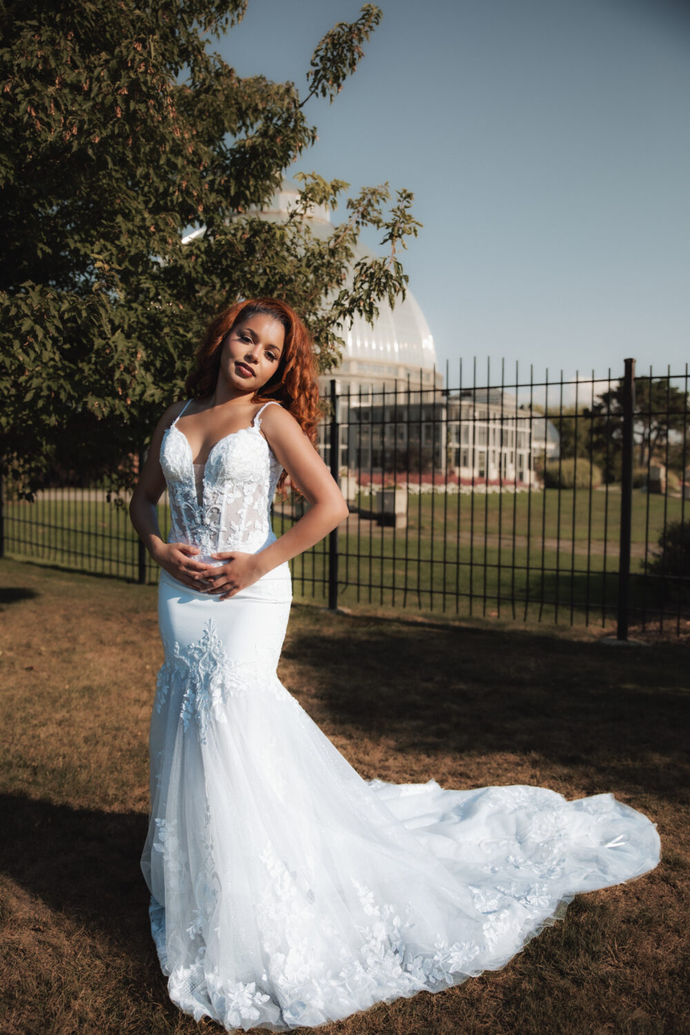 A close-up view of a bride in a lace wedding dress standing outdoors with a conservatory and trees in the background.