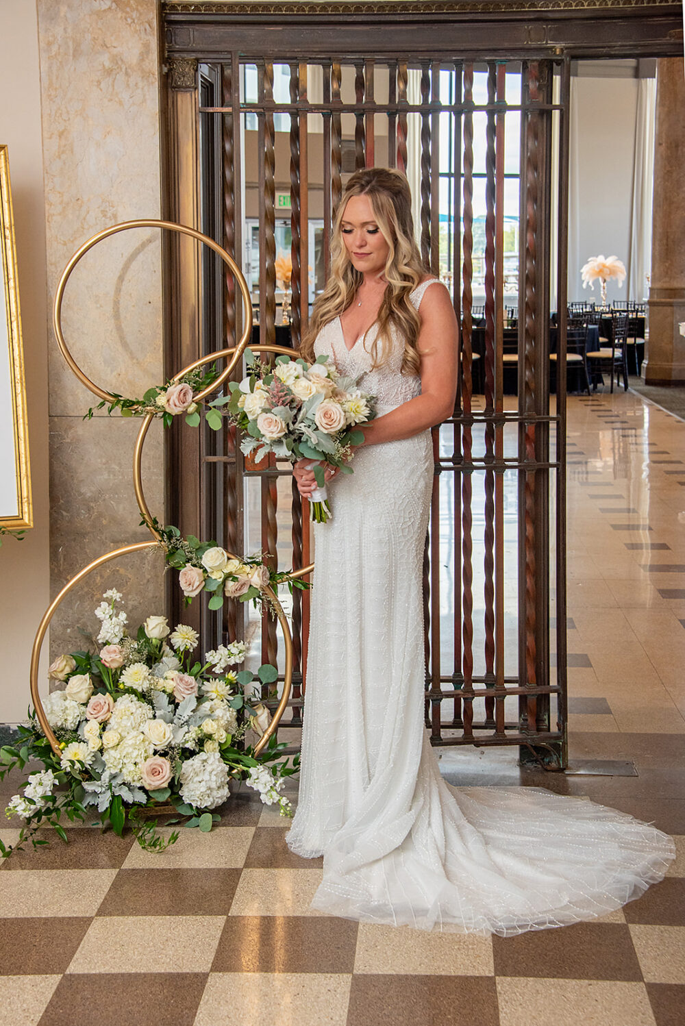 Bride in a fitted lace wedding gown holding a bouquet beside a gold circular floral display in an elegant venue hallway.