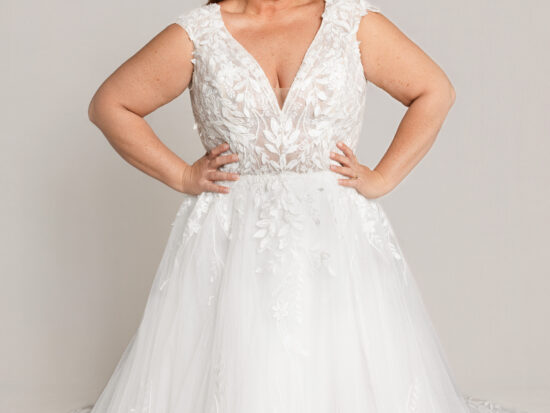 Bride wearing a sleeveless lace wedding gown with a V-neckline and full tulle skirt, standing with hands on hips.