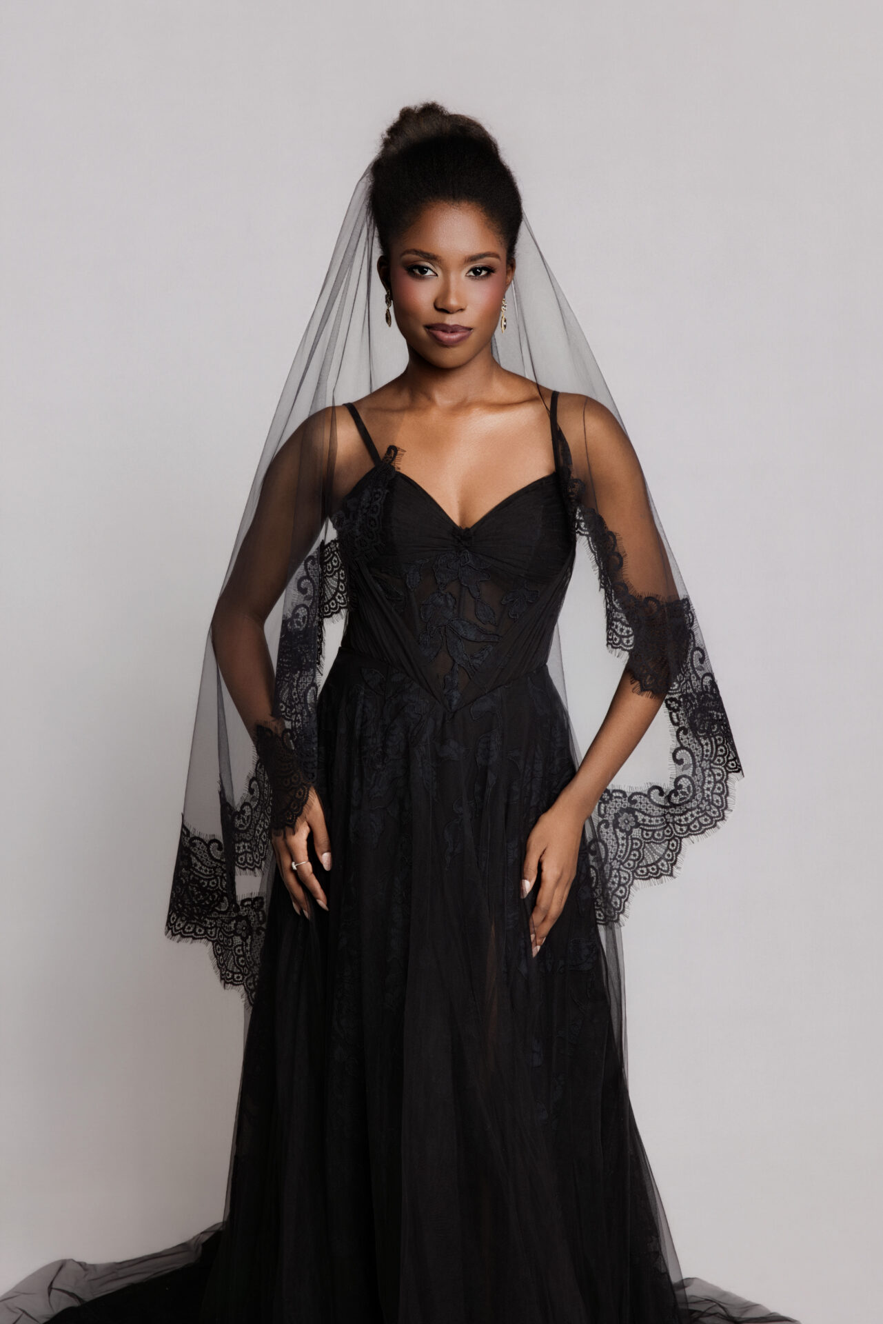 Bride wearing a black gown with sheer sleeves standing against a neutral background.