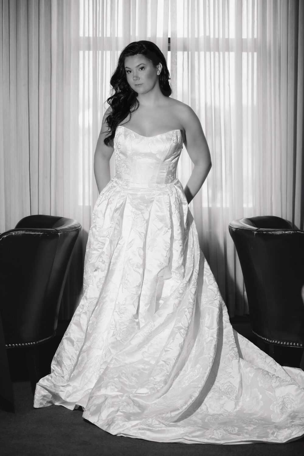 Black and white portrait of a bride in a strapless floral textured ball gown standing between two dark chairs in front of curtains.