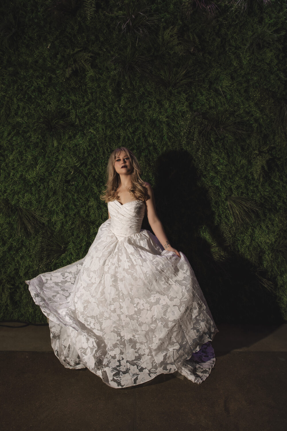 Bride in a floral textured ball gown holding up the skirt while posing in front of a green foliage wall.