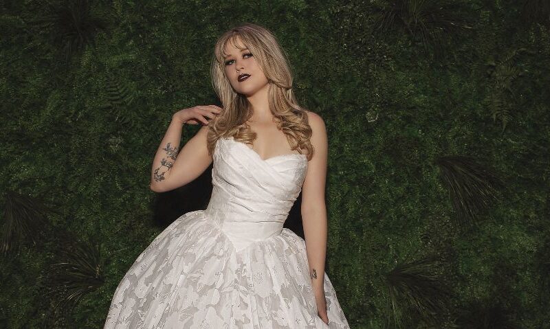 Bride in a floral textured ball gown standing in front of a green foliage wall with a chapel train spread behind her.