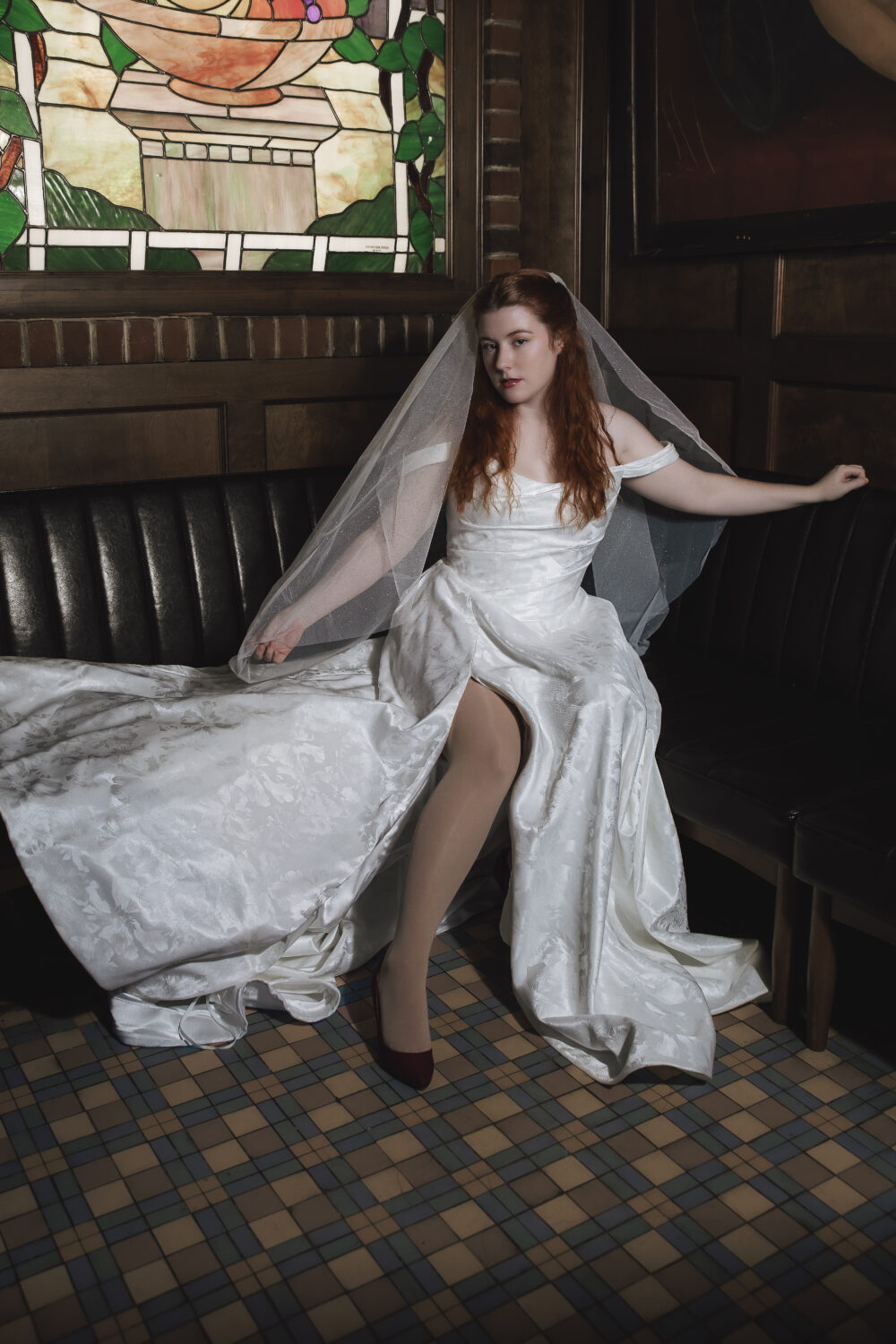 Bride in an off-the-shoulder satin wedding gown with a long veil seated on a black leather booth beneath a stained glass window.