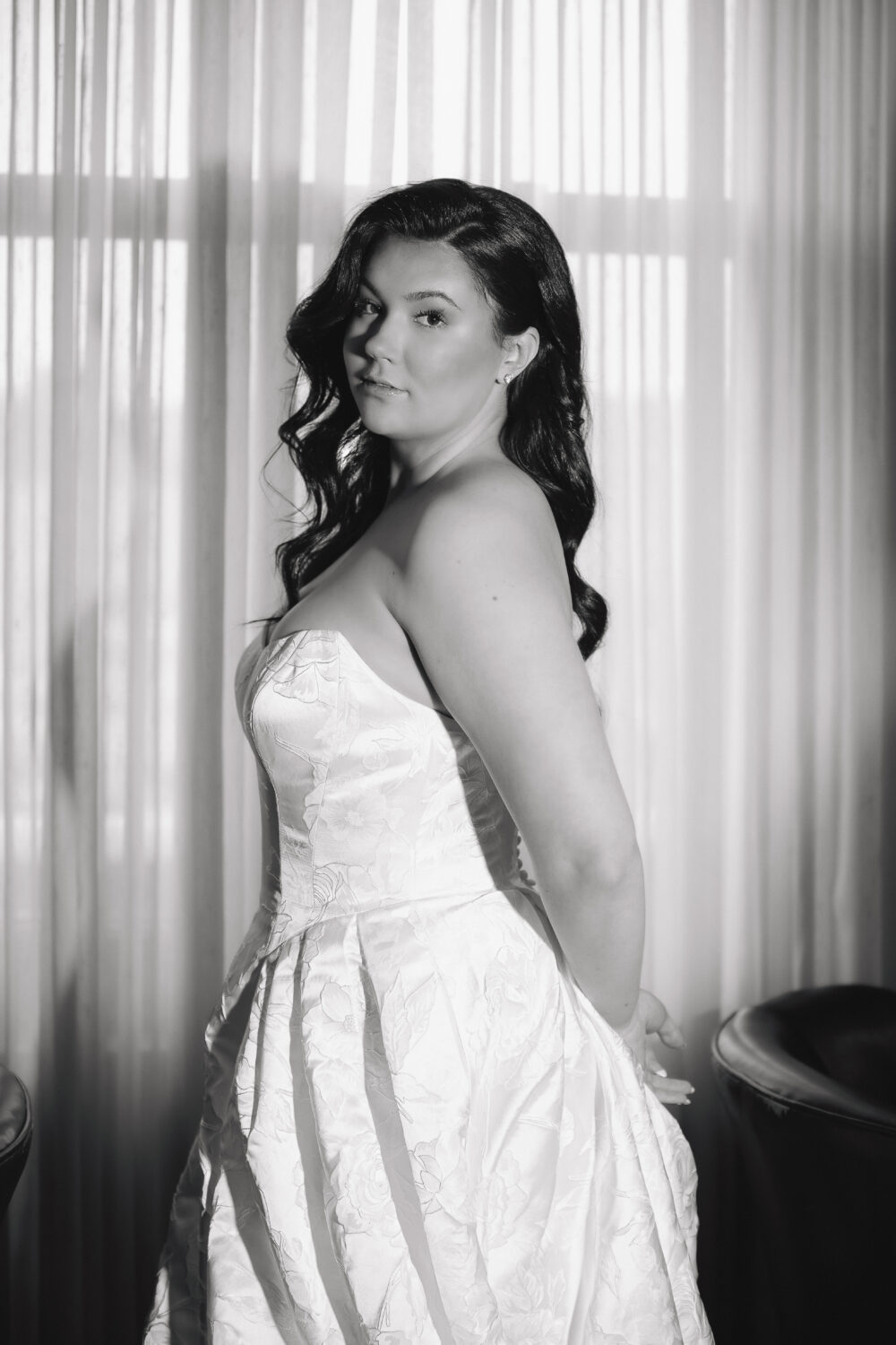 Black and white portrait of a bride in a strapless floral textured ball gown standing in front of sheer window curtains.