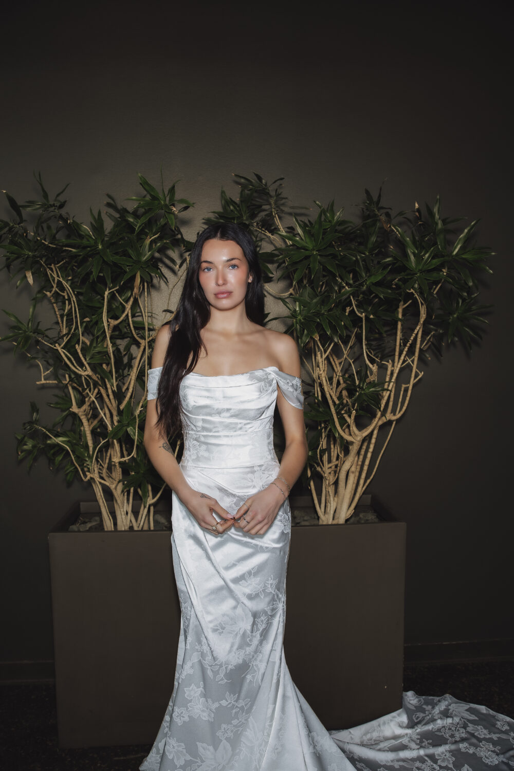 Bride in a floral textured ball gown standing in front of a green foliage wall with a chapel train spread behind her.