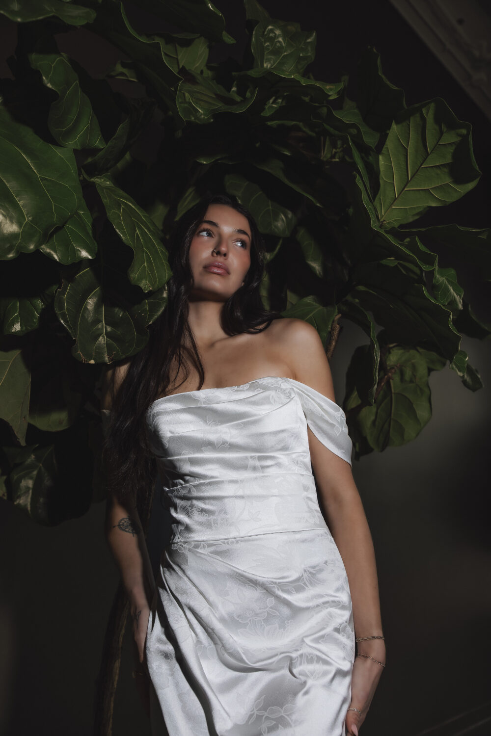 Bride in a satin off-the-shoulder wedding gown posing beside large leafy indoor plants in dramatic lighting.