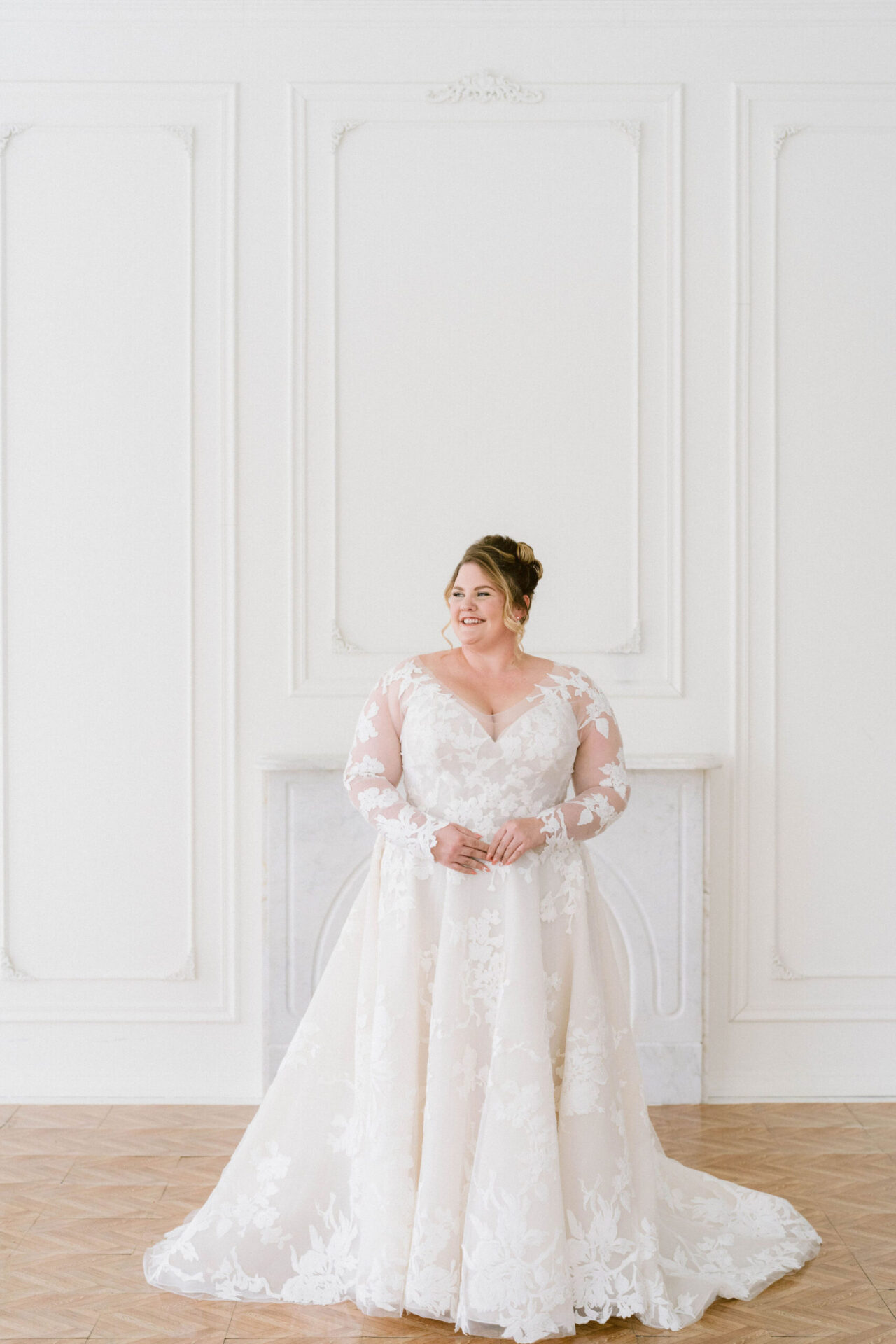 Bride wearing a long-sleeved lace wedding dress with a full skirt, standing in a classic white room.