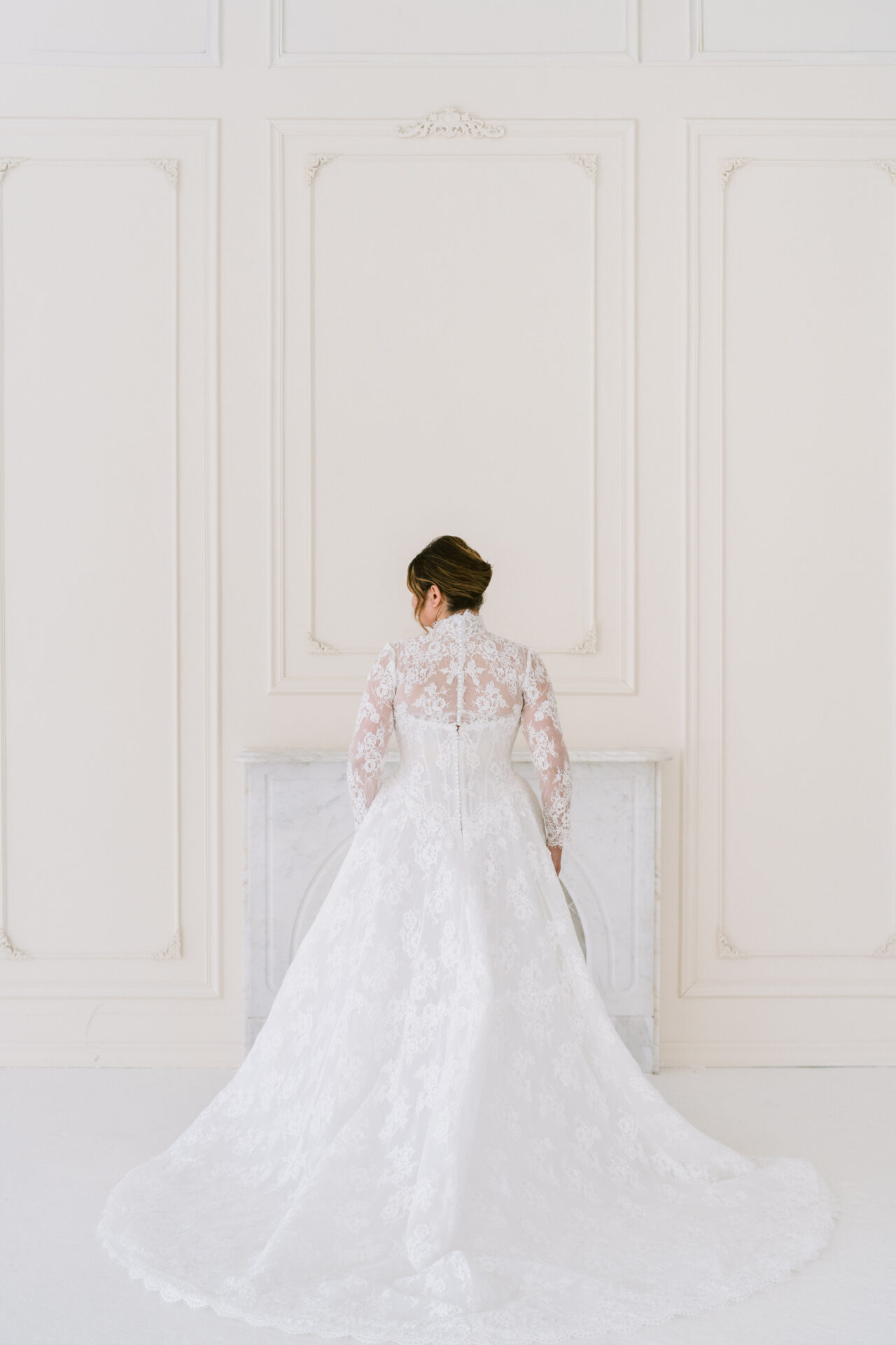 Back view of a bride in a curve lace wedding gown with long train in a white studio.
