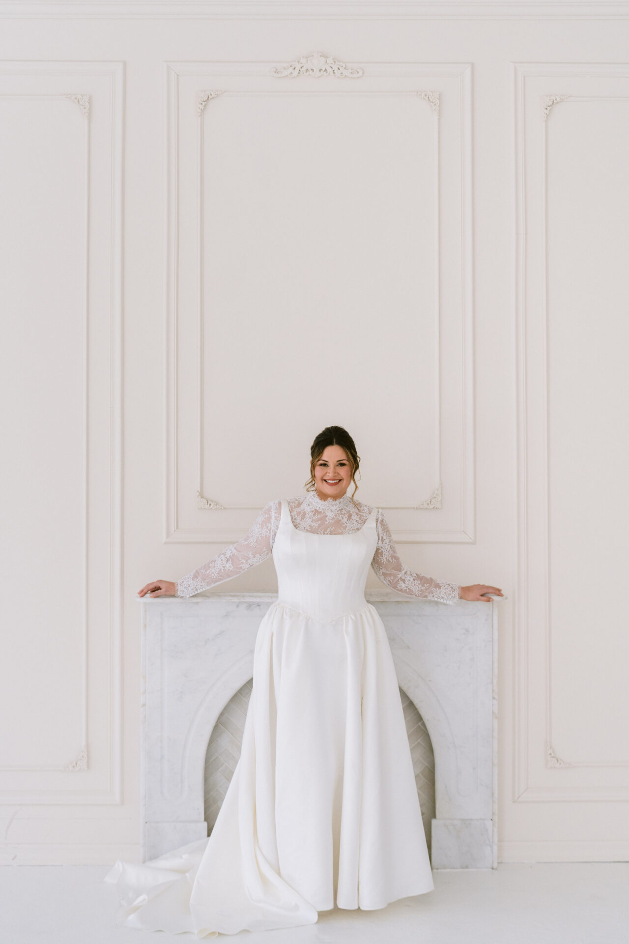 Bride in a minimalist white gown standing with arms slightly extended in a white-paneled room.