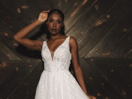 A bride in a sleeveless lace wedding dress stands against a dark wooden wall, shown in a wide cropped view.