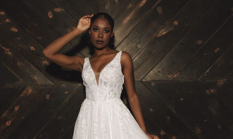 A bride in a sleeveless lace wedding dress stands against a dark wooden wall, shown in a wide cropped view.