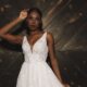 A bride in a sleeveless lace wedding dress stands against a dark wooden wall, shown in a wide cropped view.