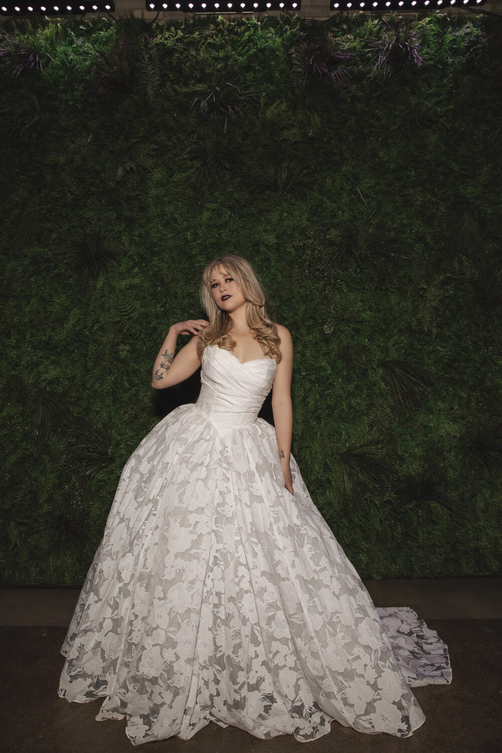 Bride in a floral textured ball gown standing in front of a green foliage wall with a chapel train spread behind her.