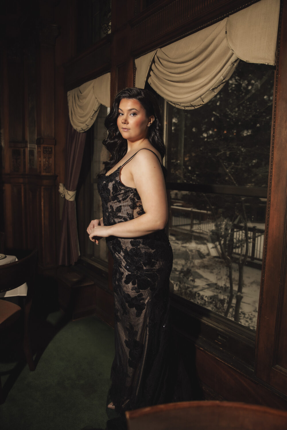 A bride in a black lace gown stands by a large window with draped curtains, overlooking a snowy outdoor scene.