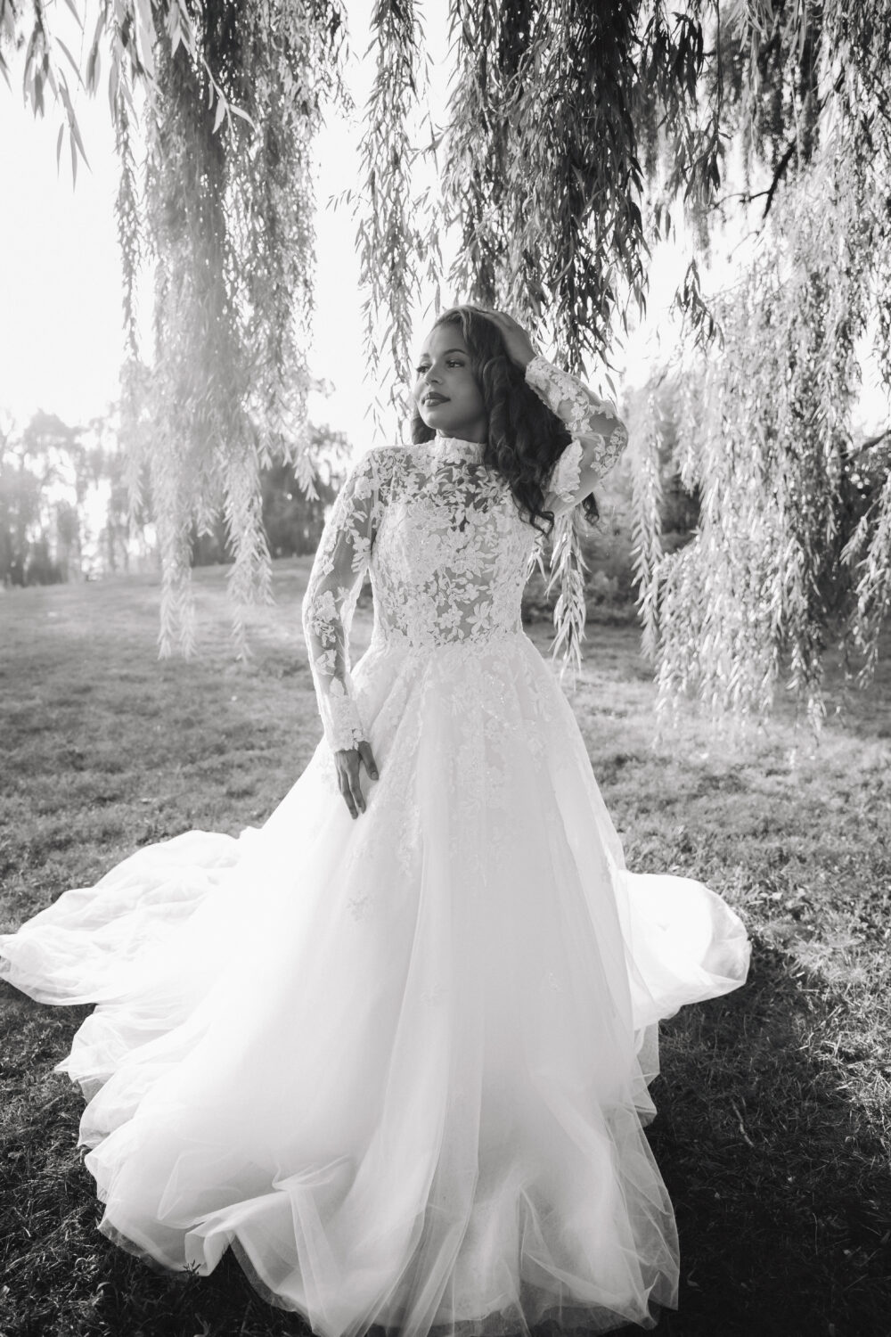 A bride in a long sleeve lace wedding dress with a high neckline stands outdoors under hanging tree branches in a black-and-white photo.