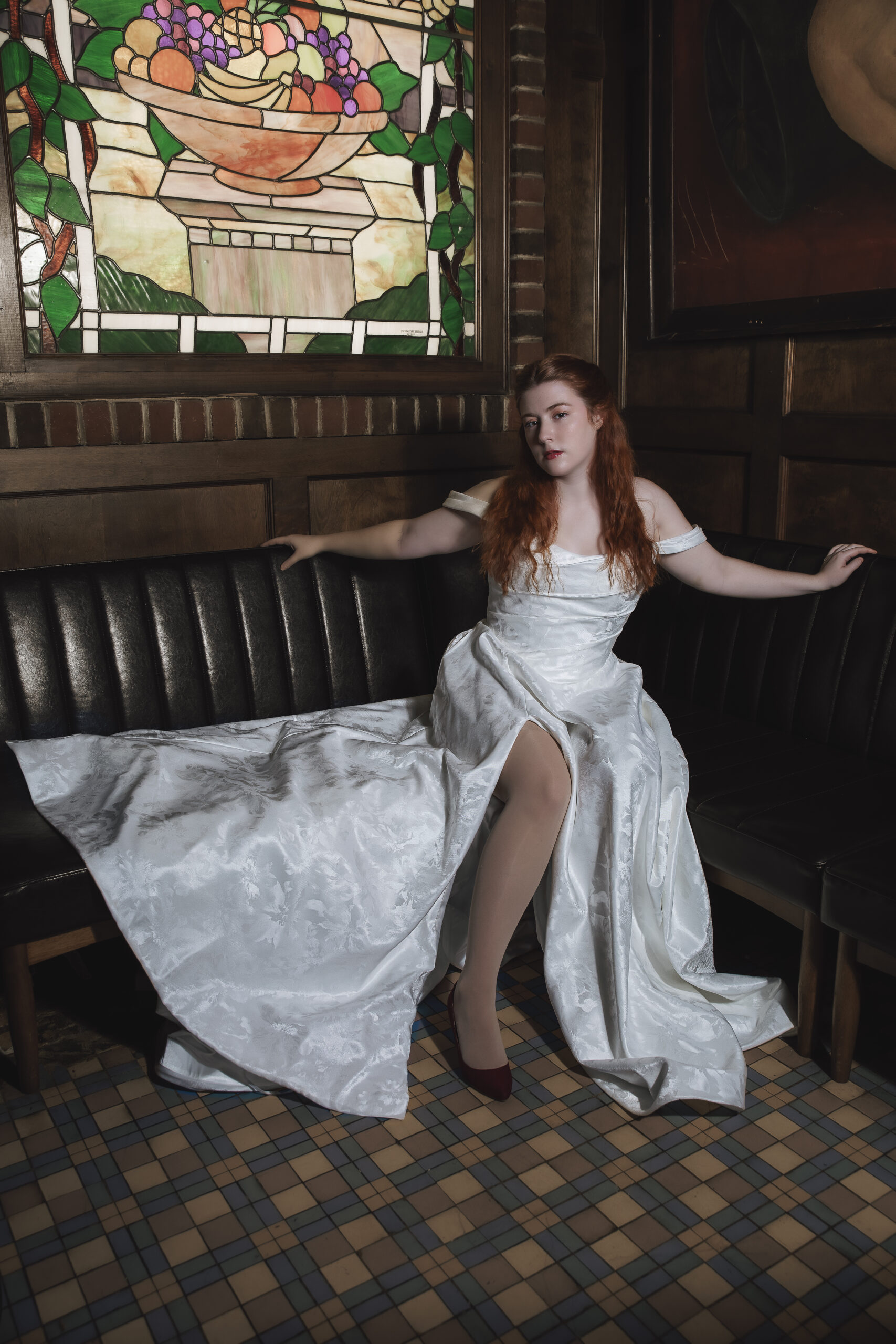 Bride in an off-the-shoulder satin gown seated on a black leather booth with a stained glass window in the background.