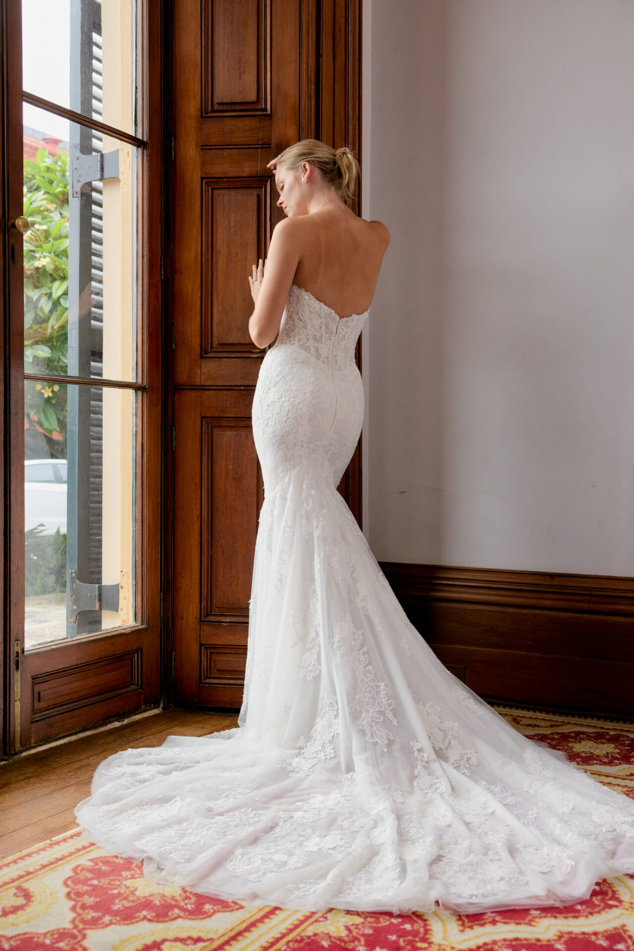 Bride in a fitted lace gown standing near a window with a long train.