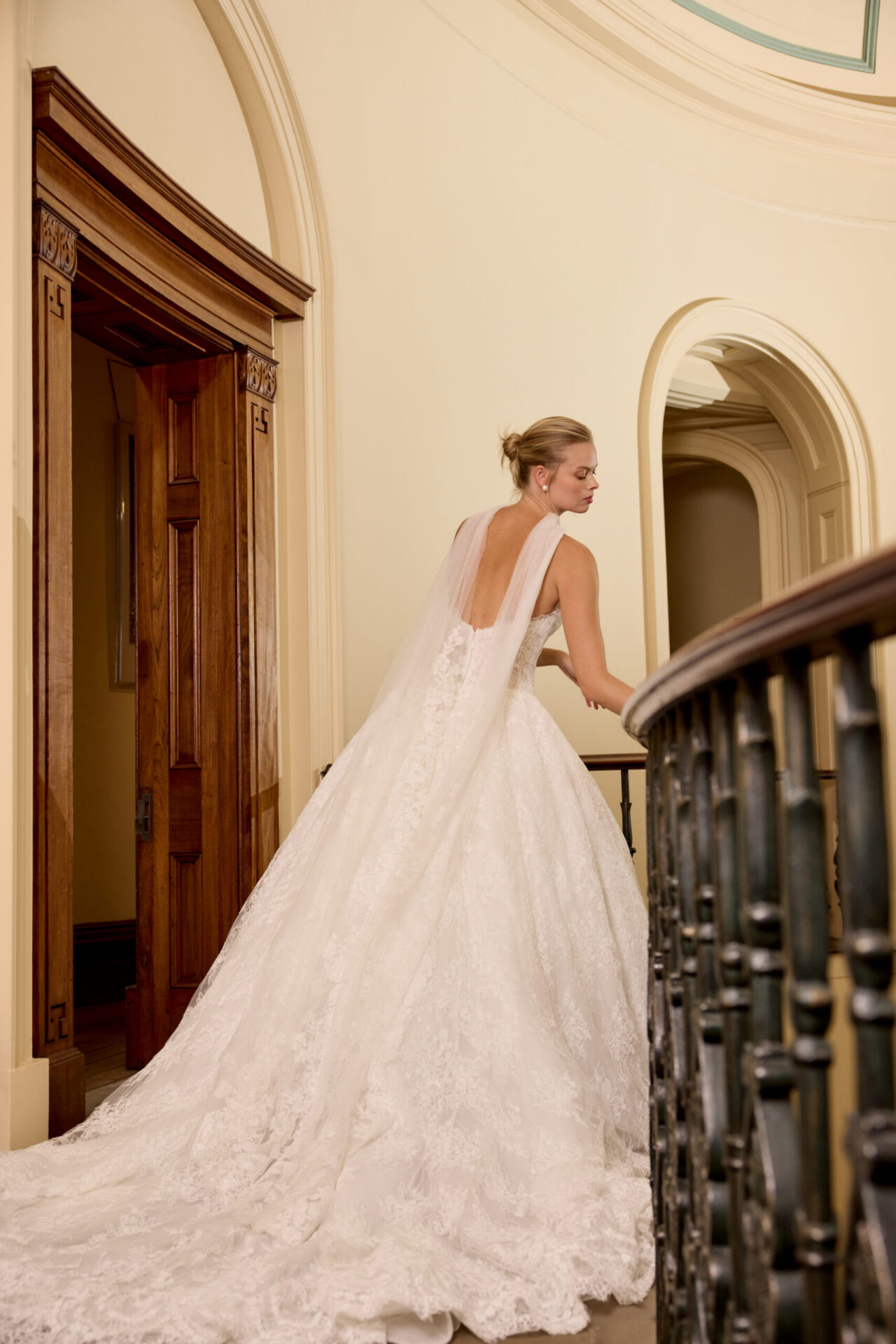 Bride in a strapless lace ball gown walking along an indoor balcony with ornate railing.