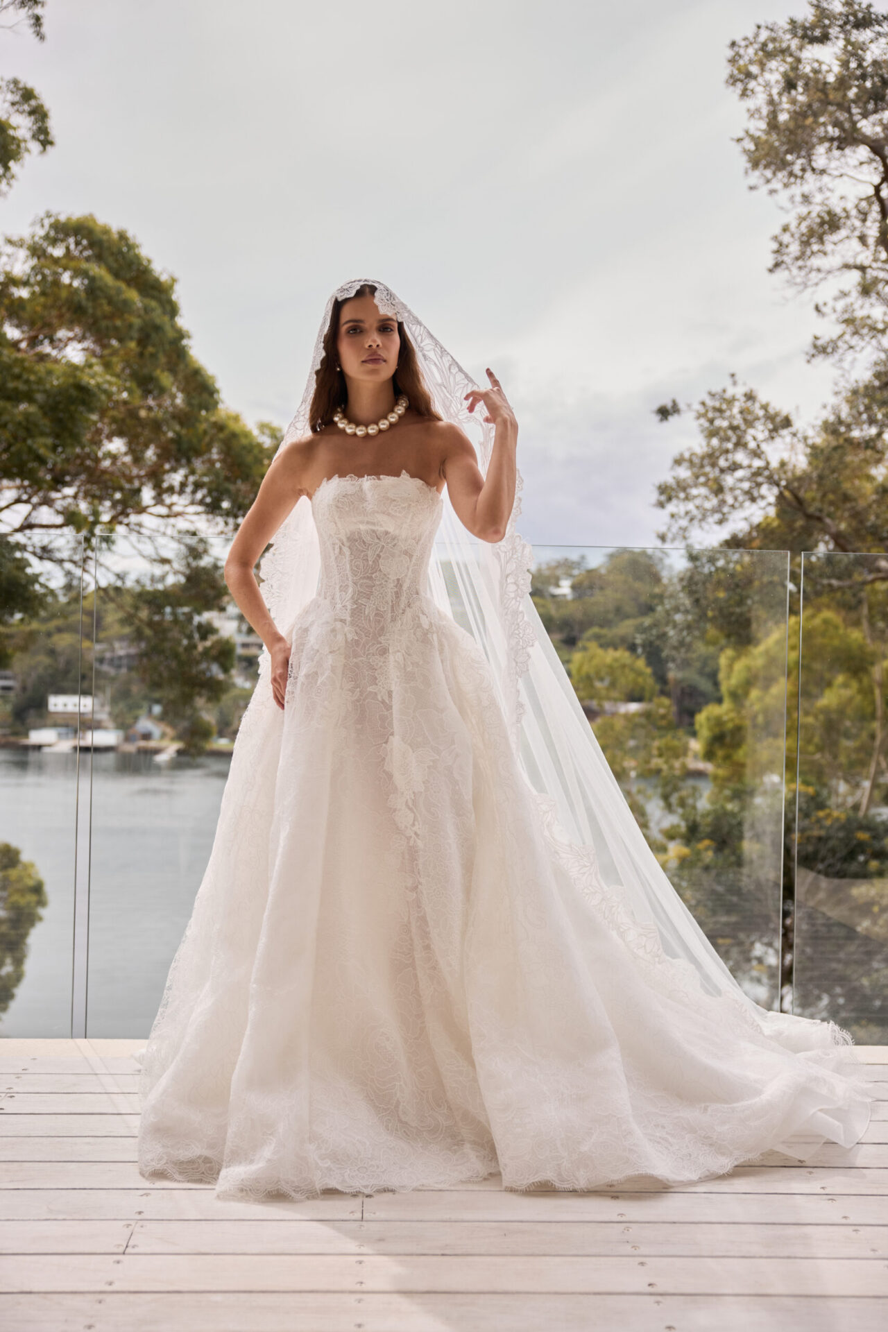 Bride in a strapless ball gown standing outdoors near water and trees.
