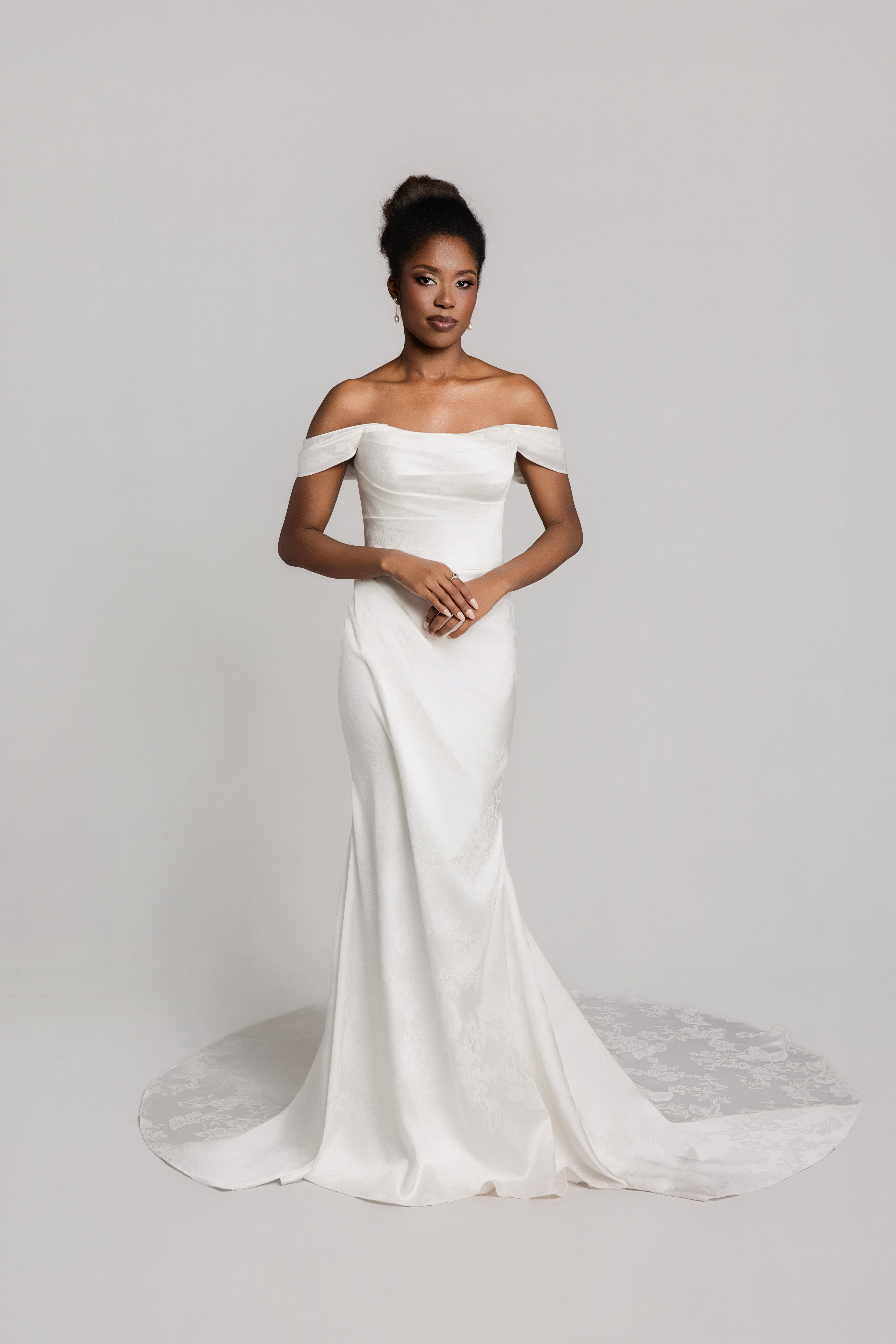Bride wearing an off-the-shoulder fitted white wedding gown with a mermaid silhouette and slight train, standing against a neutral background.