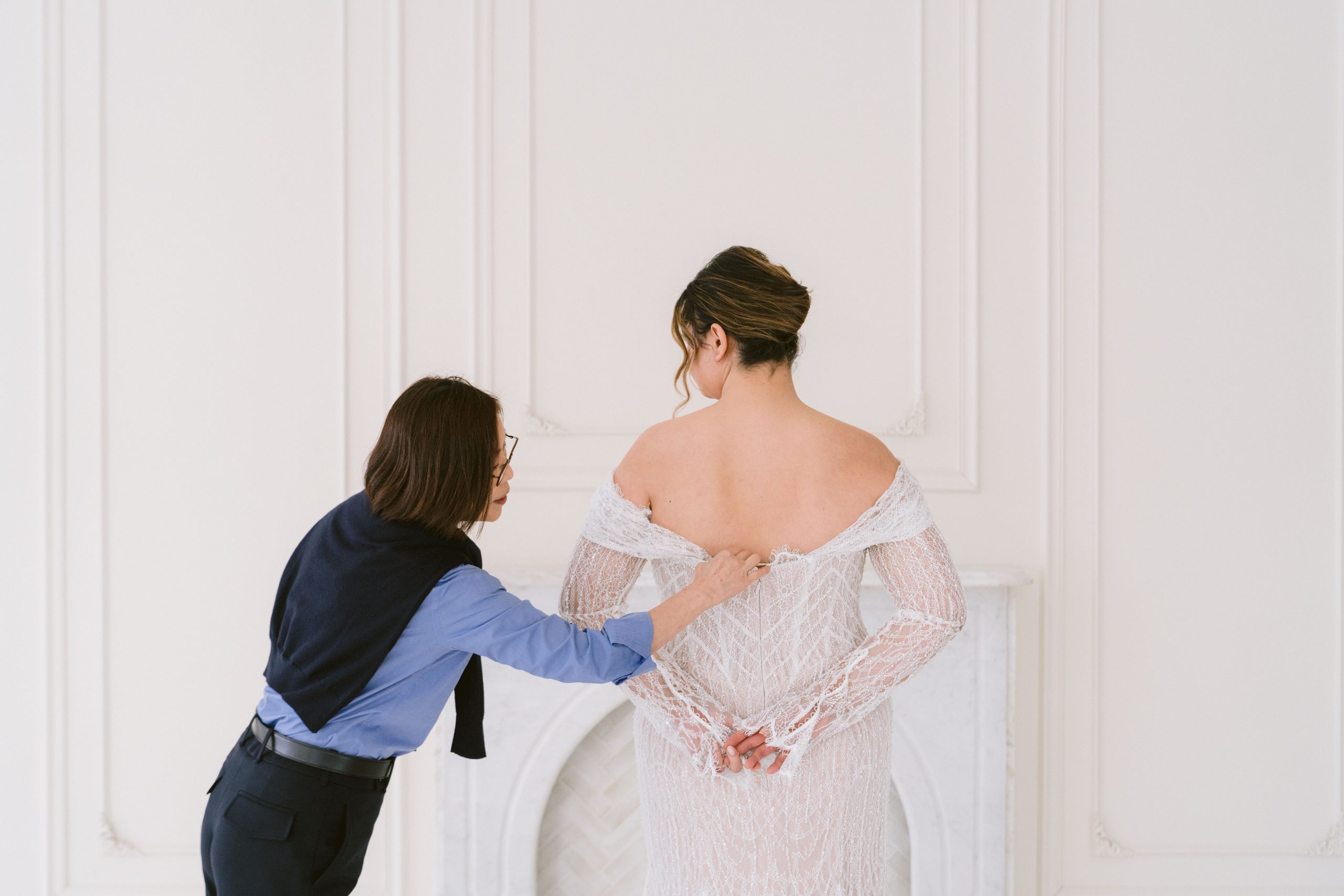 Assistant adjusting the back of a bride’s off-shoulder lace wedding dress in a studio setting.