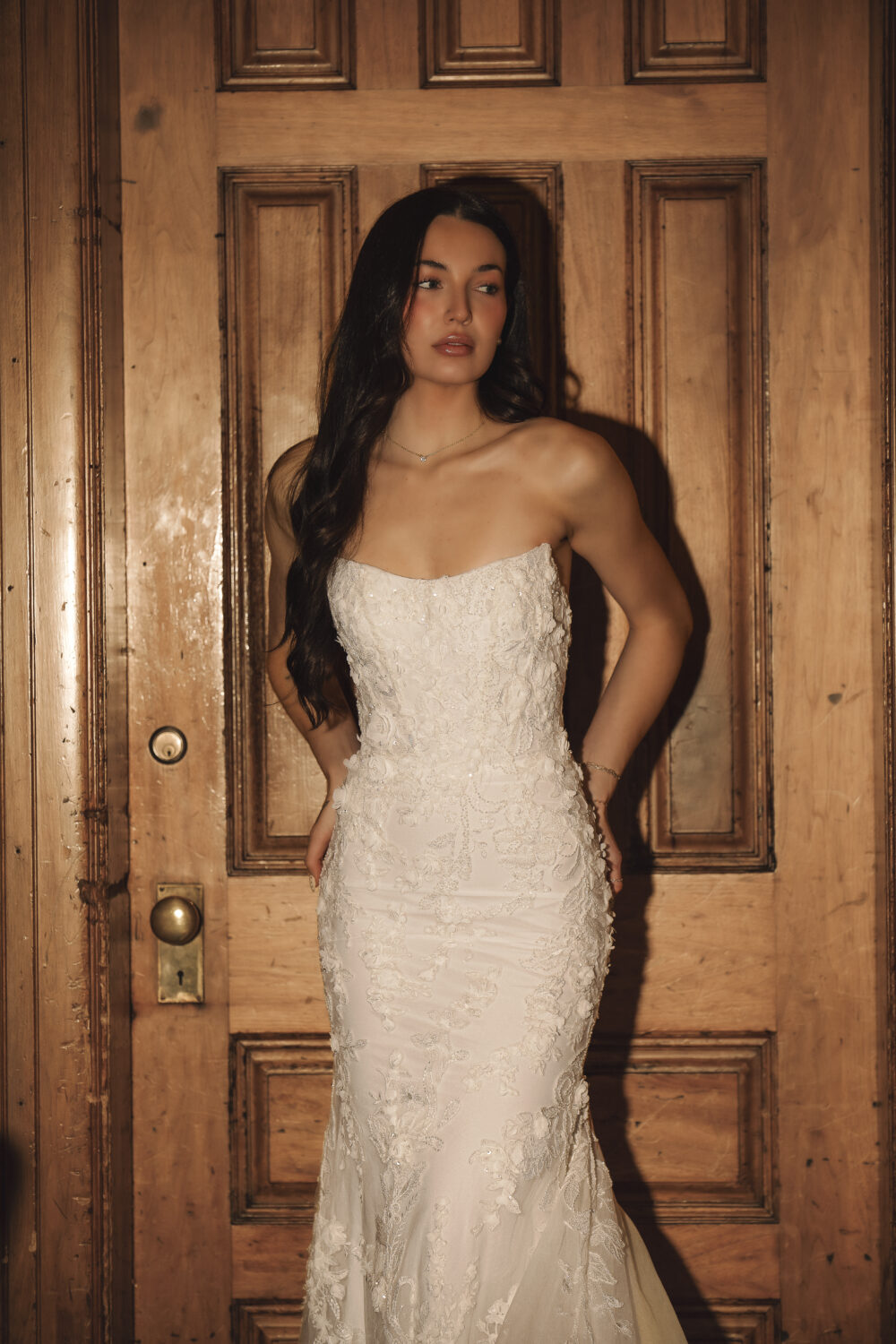 A bride in a strapless lace wedding dress stands against a wooden door, looking to the side in warm indoor lighting.