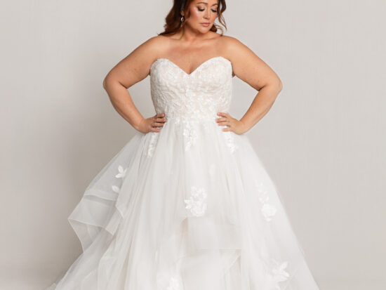 Bride wearing a strapless white wedding gown with a sweetheart neckline and layered tulle skirt, standing with hands on hips against a neutral background.