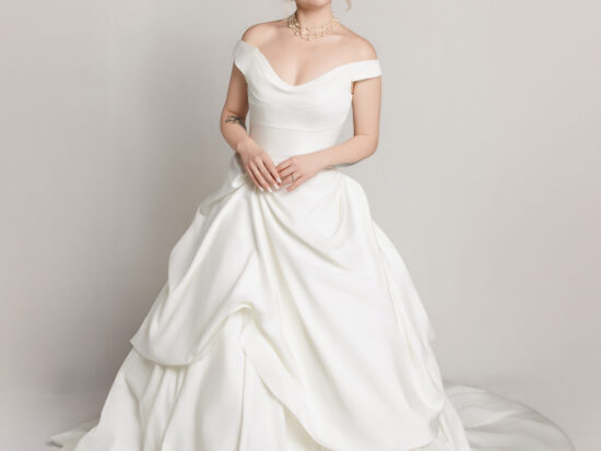 Bride in an off-the-shoulder white wedding gown with a full skirt and soft draping, seated slightly and posing against a neutral background.