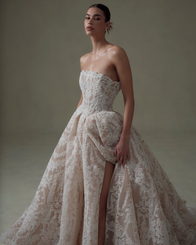 Bride wearing a strapless lace ball gown with a fitted bodice and high slit, posing against a neutral background.