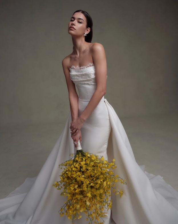 Bride in a strapless white gown holding a bouquet of yellow flowers, posing against a neutral studio background.