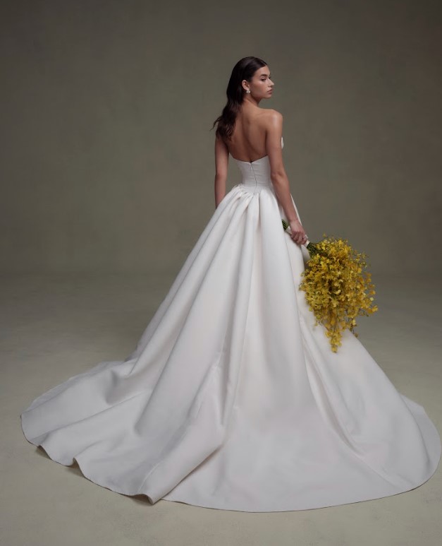 Back view of a bride in a strapless white gown with a long train, holding a bouquet of yellow flowers against a neutral background.