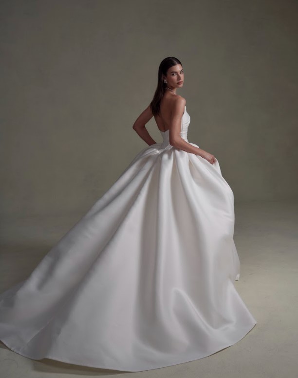 Back view of a bride in a strapless white ball gown with a voluminous skirt, looking over her shoulder against a neutral background.