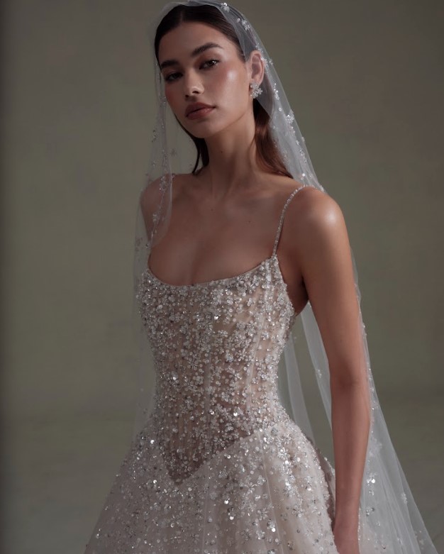 Close-up of a bride wearing a sparkling beaded wedding gown with thin straps and a sheer veil against a neutral background.