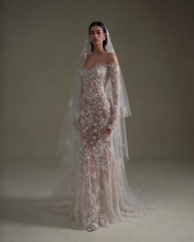 Bride wearing a sheer lace wedding gown with long sleeves and a veil, standing against a neutral studio background.