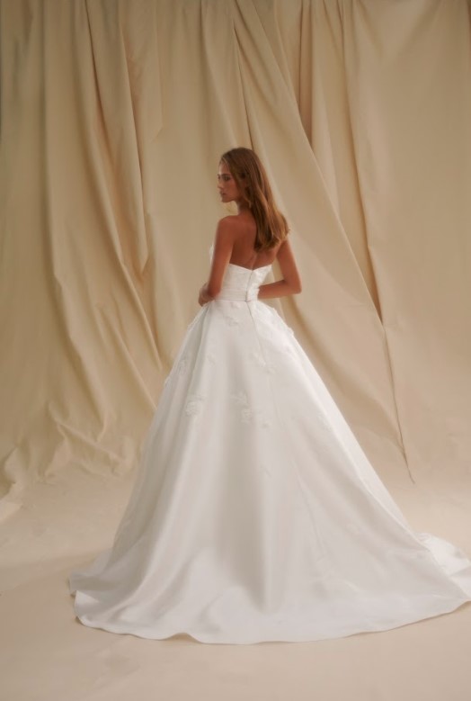 Bride in a strapless white wedding gown with a full flowing skirt, shown in side profile against a draped neutral backdrop.