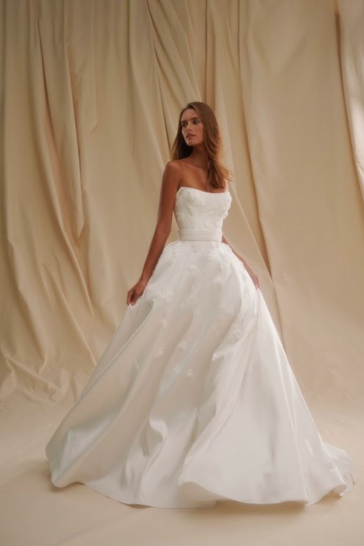 Bride wearing a strapless white ball gown walking forward, holding part of the skirt against a soft draped background.