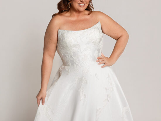 Close-up of a smiling bride in a one-shoulder white wedding gown with floral detailing, posing with one hand on her hip.