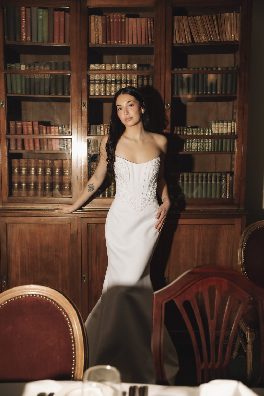 A bride in a fitted strapless wedding dress stands in front of a wooden bookshelf filled with books inside a vintage dining room.