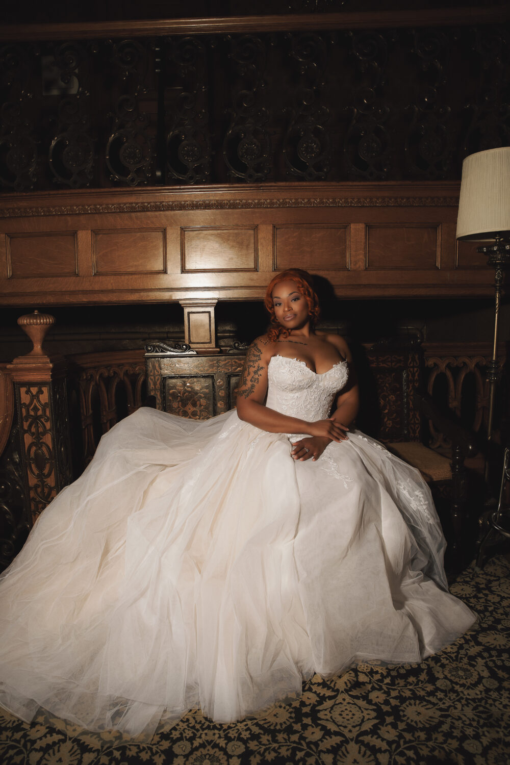A bride in a voluminous ball gown wedding dress sits in a dimly lit, historic interior.