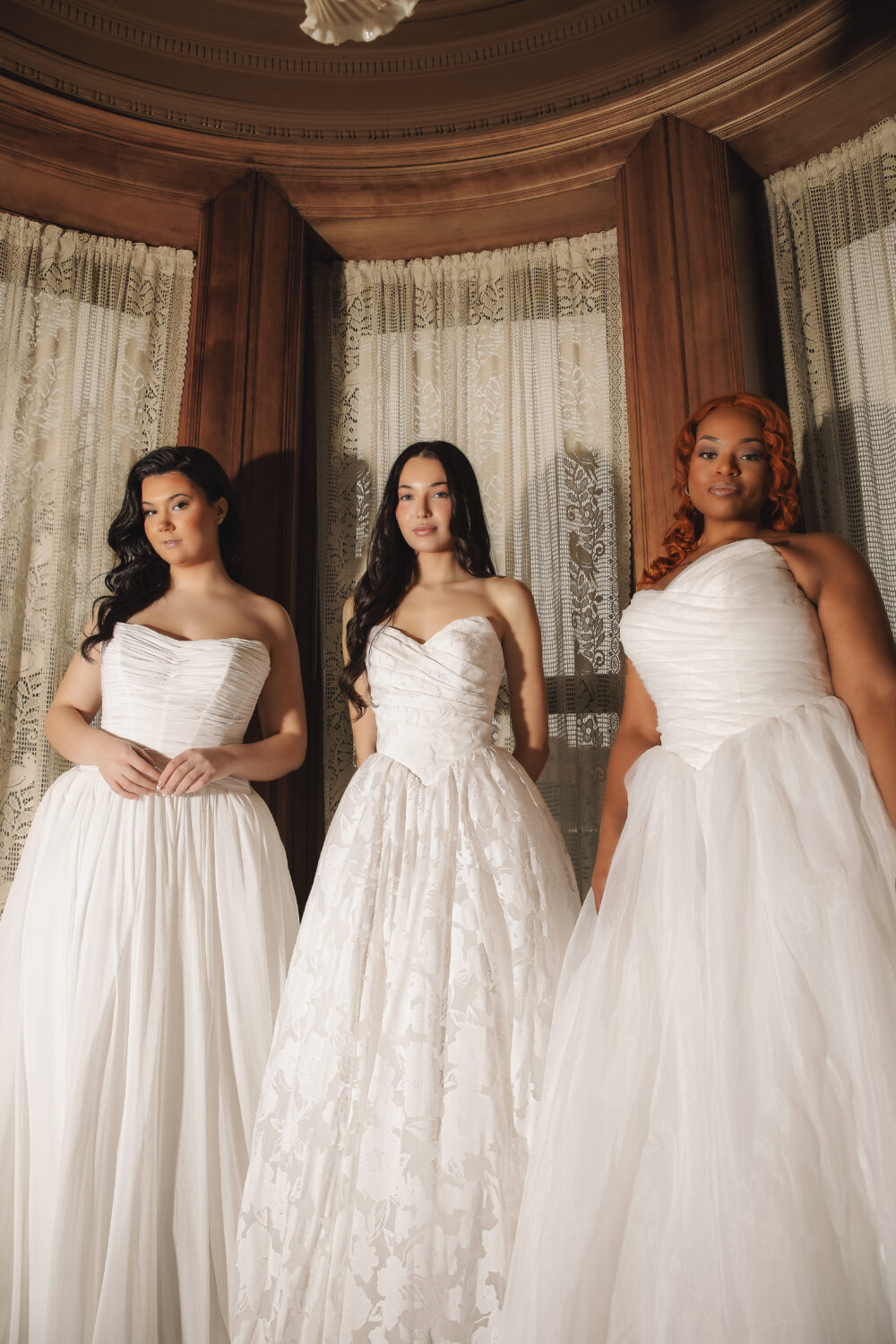 Three brides stand side by side wearing strapless and one-shoulder wedding dresses in a softly lit room with lace curtains.
