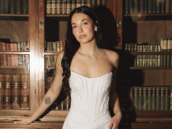 A bride in a fitted strapless wedding dress stands in front of a wooden bookshelf filled with books inside a vintage dining room.
