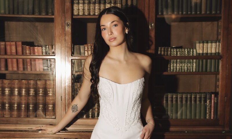 A bride in a fitted strapless wedding dress stands in front of a wooden bookshelf filled with books inside a vintage dining room.