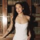 A bride in a fitted strapless wedding dress stands in front of a wooden bookshelf filled with books inside a vintage dining room.
