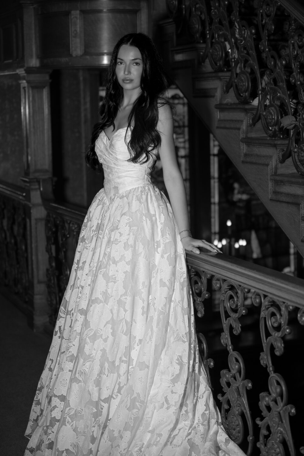 A bride in a strapless floral wedding dress leans on a decorative railing near an ornate staircase in a black-and-white image.