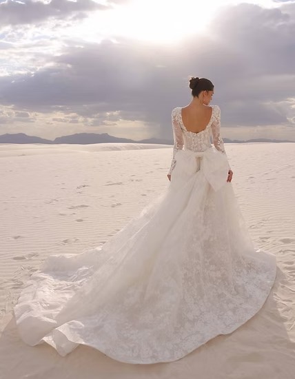 Back view of a bride in a lace wedding gown walking through white sand dunes with a long train trailing behind.