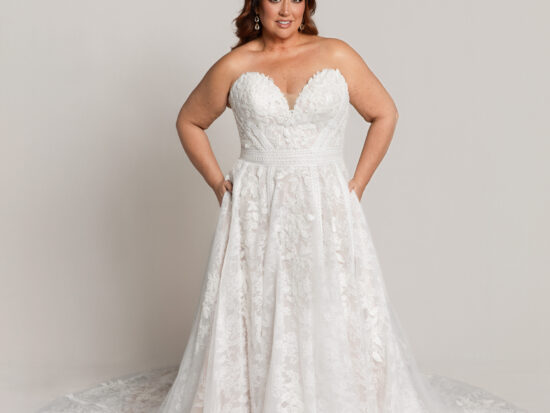 Bride wearing a strapless lace wedding gown with a sweetheart neckline and full skirt, standing with hands on hips.