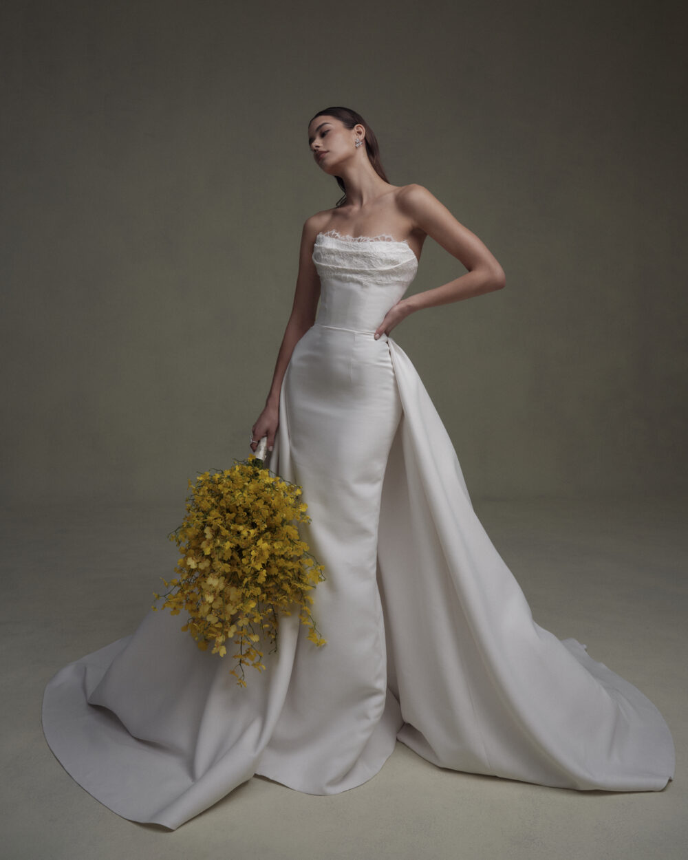 Model wearing a fitted strapless wedding gown with a long overskirt and holding a bouquet of yellow flowers.