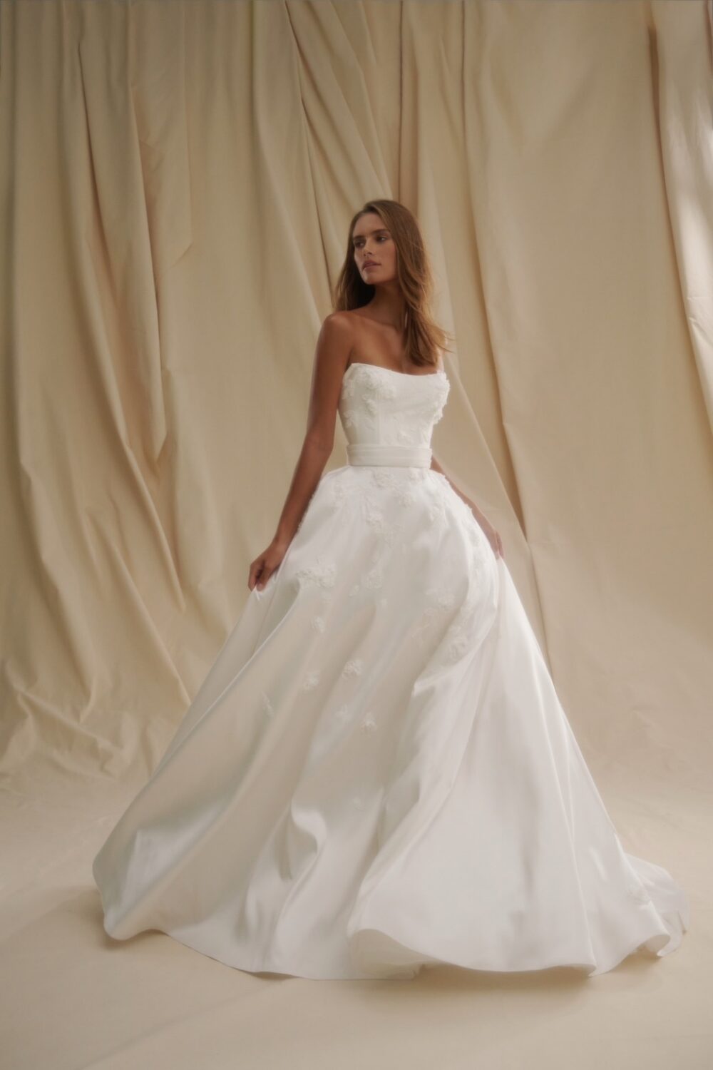 Model in a strapless ball gown wedding dress with floral detailing posed in front of draped fabric panels.
