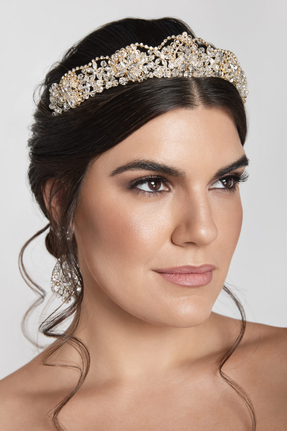 A close-up portrait of a woman with an elegant updo wearing a gold and crystal tiara and statement earrings against a neutral background.